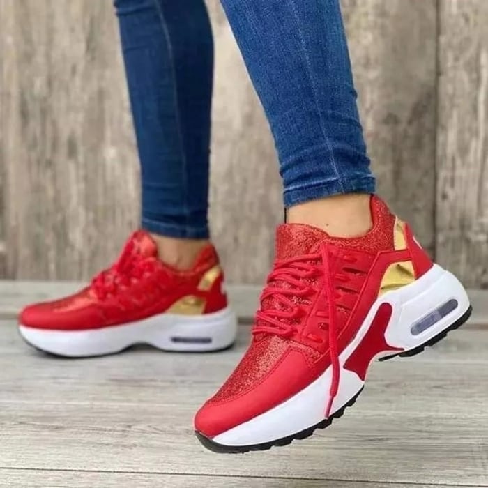 Red women's sneakers with gold accents, white sole, worn by a person in blue jeans. Fashionable sneakers, ideal for leisure and sport.